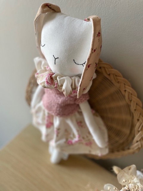 Doudou fait main Isaure en coton recyclé, portant une jupe fleurie en tissu vintage, un pull rose poudré et un col romantique, dans un décor naturel et chaleureux.