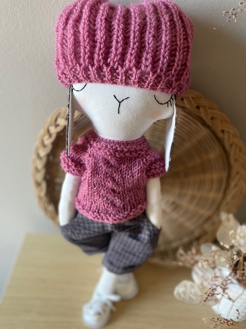 Lou, un doudou artisanal en coton recyclé, portant un pull rose poudré, un bonnet assorti et un baggy gris foncé