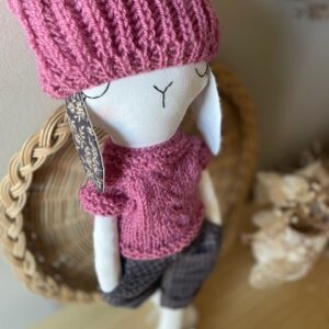 Lou, un doudou artisanal en coton recyclé, portant un pull rose poudré, un bonnet assorti et un baggy gris foncé